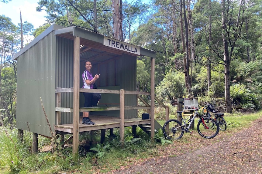 A woman in bike-riding gear stands and smiles from a shack in a Tasmanian forest, with several mountain bikes nearby.