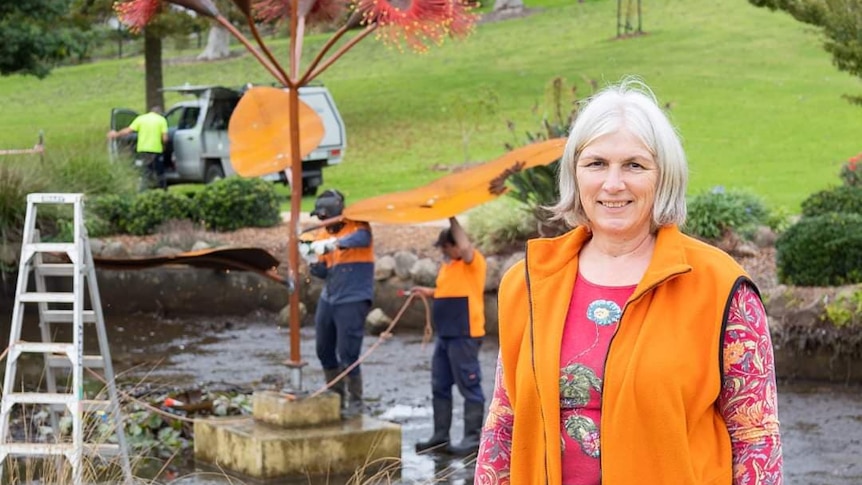 Jo in a bright orange vest in a garden as a workers tend to a sculpture in the background. 