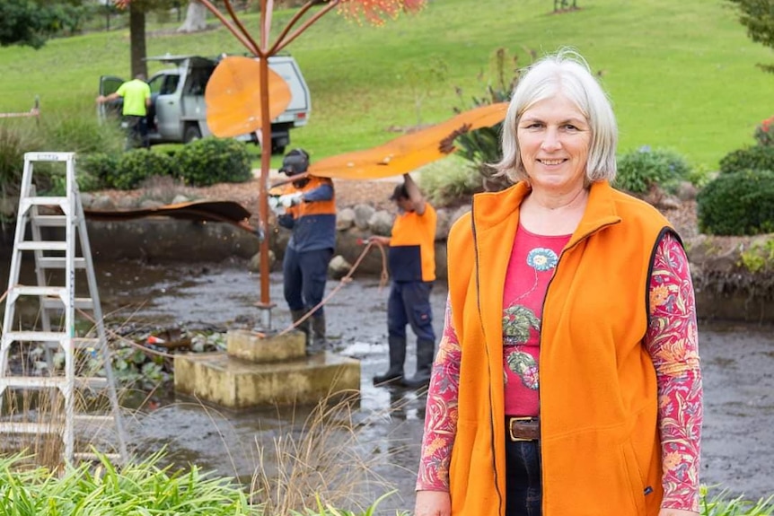 Jo in a bright orange vest in a garden as a workers tend to a sculpture in the background. 
