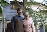 Man and woman smiling in front of their weatherboard home painted blue and cream, near a tree.