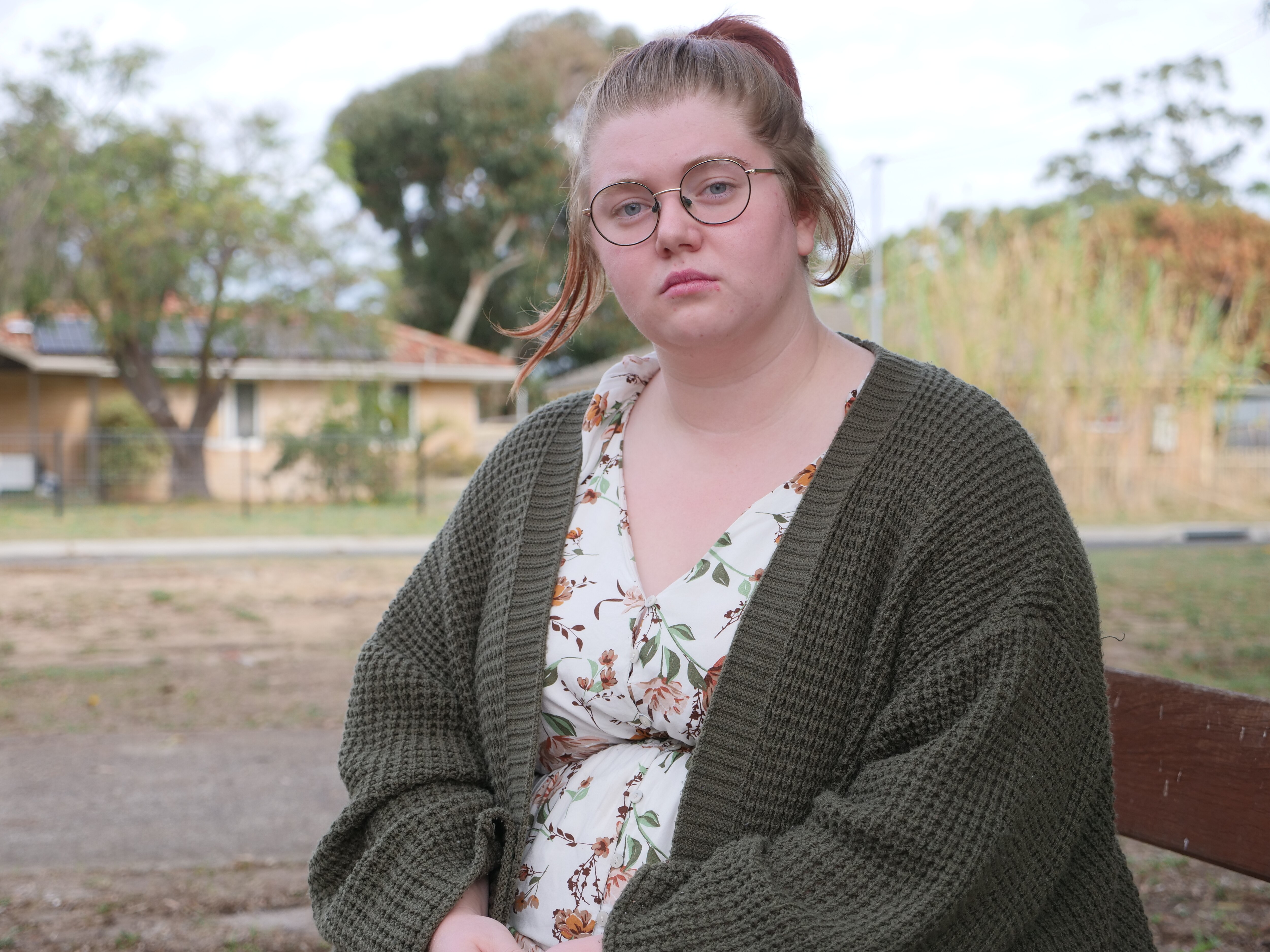 A woman in dark green cardigan and white top looks neutrally at the camera. It is overcast, trees in the background.