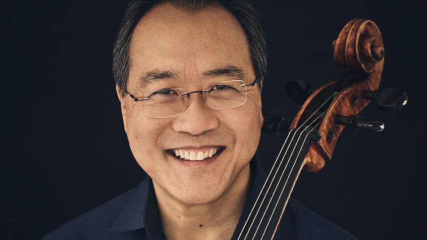 Yo-Yo Ma holds a cello and looks directly at the camera smiling. He wears a navy shirt.