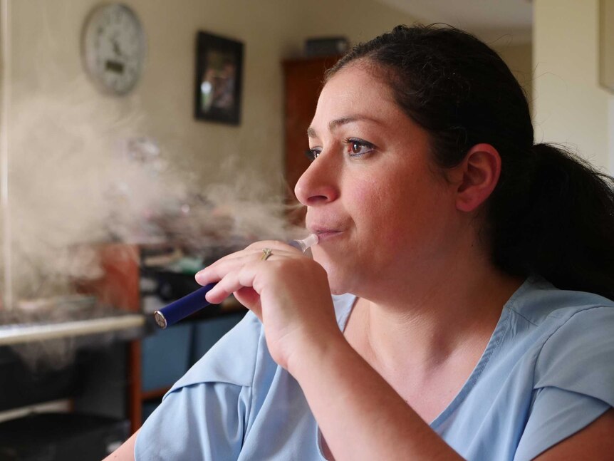 A woman with brown hair in a pony tail exhaling from a vape with the gas in front of her