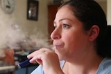 A woman with brown hair in a pony tail exhaling from a vape with the gas in front of her