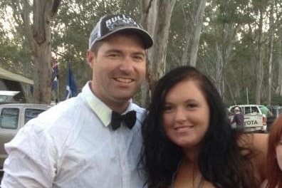 A man in a cap and a bow tie with a white shirt puts his arm around a woman with a black dress and hair. They are in the woods.