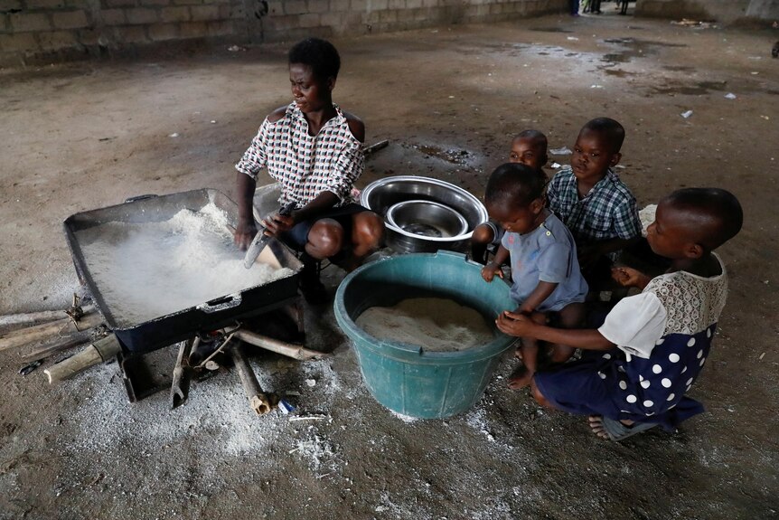 A woman prepares white powder in a large pan on a dirt floor as young children look on.