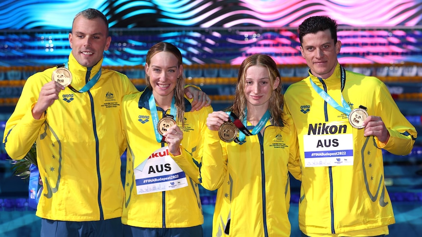 Swimmers wearing yellow jackets hold up medals and pose for a photo after winning a race
