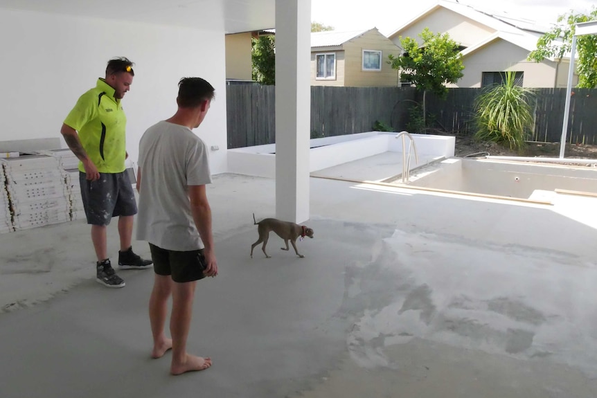 Two men look at an empty pool and a small dog walks in the shot