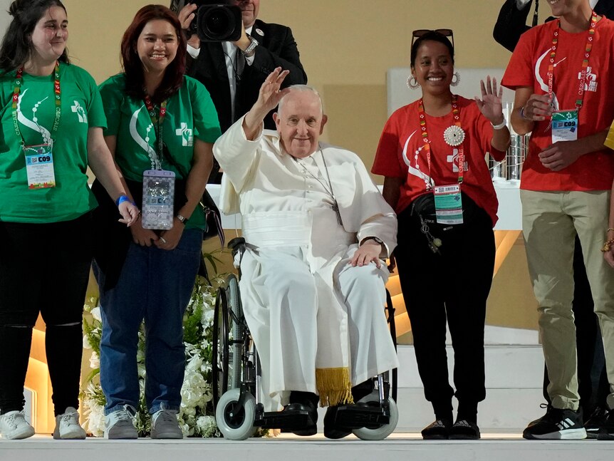A pope in a wheelchair with people standing on his right and left.