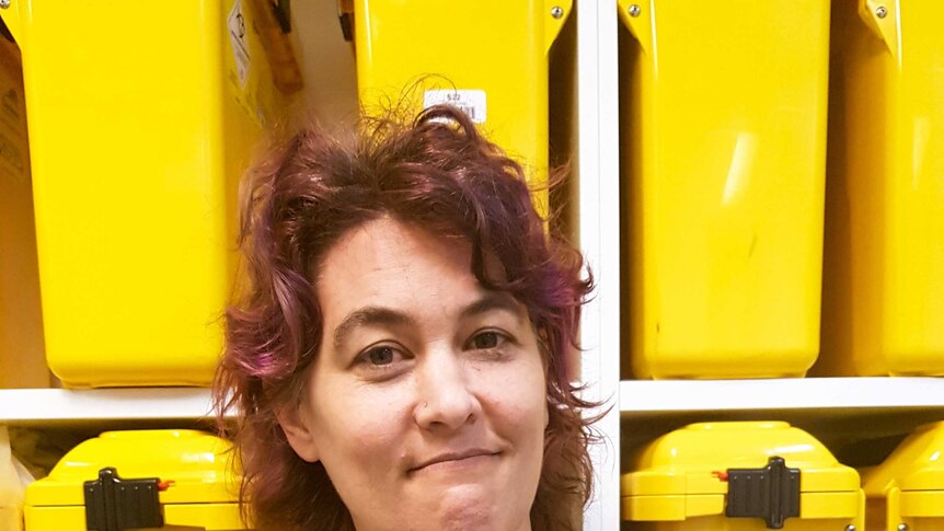 Dr Marianne Jauncey standing in front of yellow containers on a shelf holding a needle
