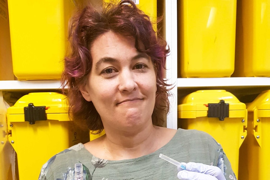 Dr Marianne Jauncey standing in front of yellow containers on a shelf holding a needle