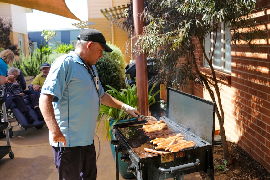 A man wearing a blue shirt and black cap turns sausages on a barbecue.