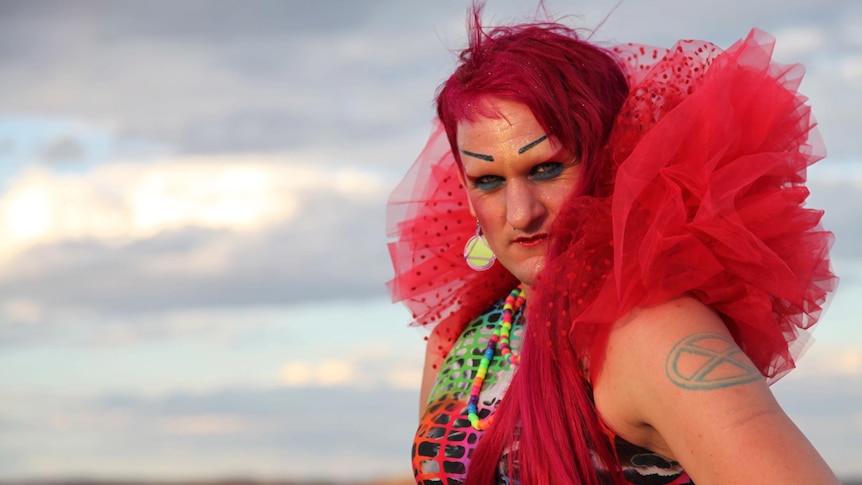 A woman with pink hair poses and looks to the camera as she stands in a field. She wears a pink tulle ruffle.