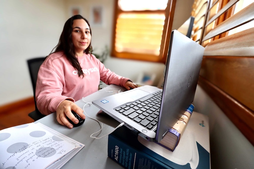 A young woman sits at a desk in front of a laptop, which is raised higher on books, working from her home office