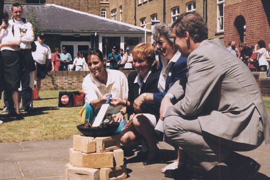 Belinda Clark holds a bat over a small fire pit, with two women and a man kneeling beside her.