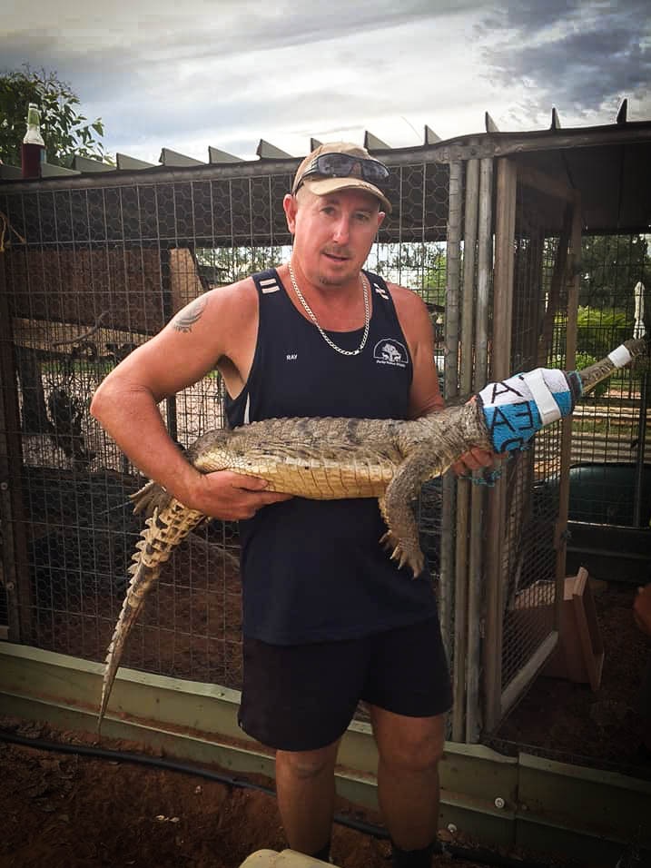 A man holds a small crocodile with its face wrapped in material. He stands in front of an enclosure.