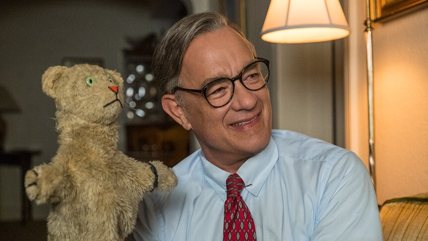 An older man in shirt and tie with greying hair and glasses sits on couch and smiles while controlling brown tiger puppet.