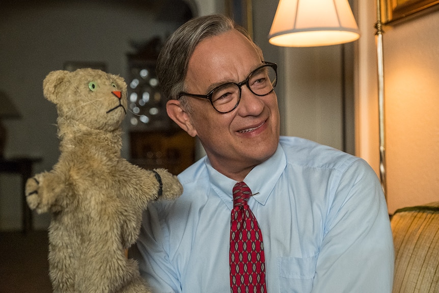 An older man in shirt and tie with greying hair and glasses sits on couch and smiles while controlling brown tiger puppet.