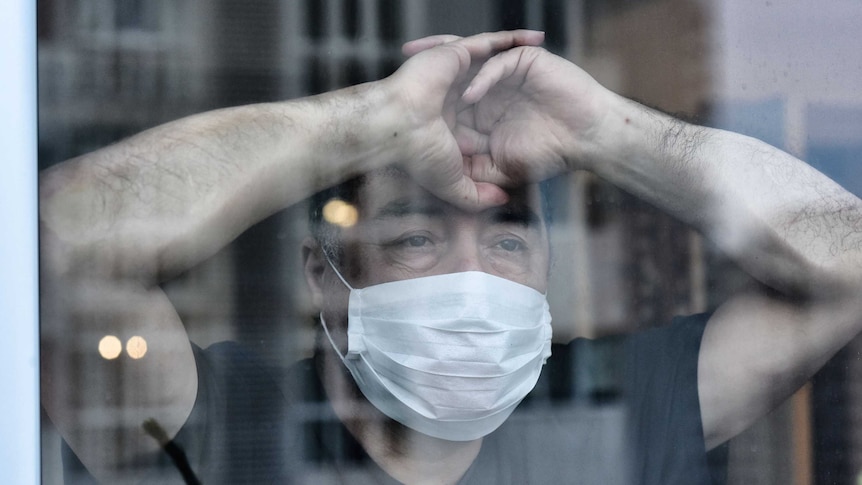 Man wearing face mask and staring out of a closed window looking sad