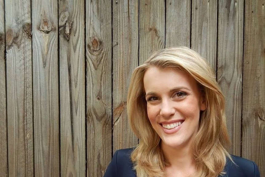 Blonde woman in blue jacket standing against fence.