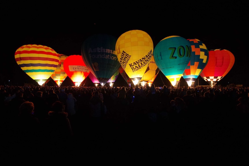 Hot air balloons light up the night sky, with thousands of people silhouetted by the bright light.