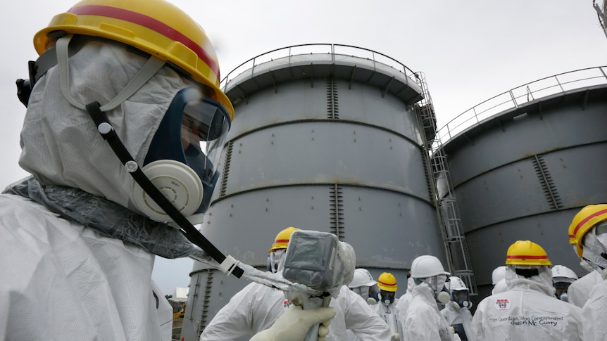 A group of men in white hazard suits, masks and yellow helmets stand beside a group of large, green-grey water tanks.