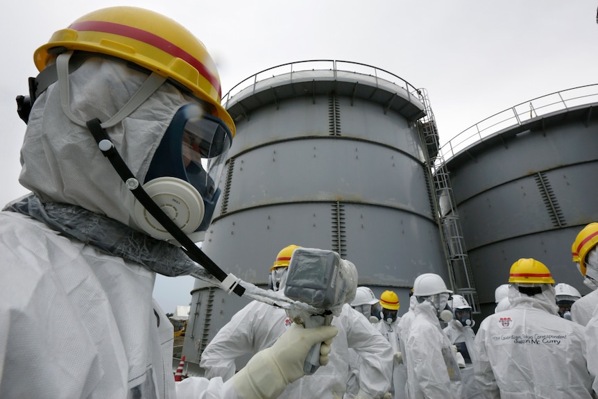 A group of men in white hazard suits, masks and yellow helmets stand beside a group of large, green-grey water tanks.