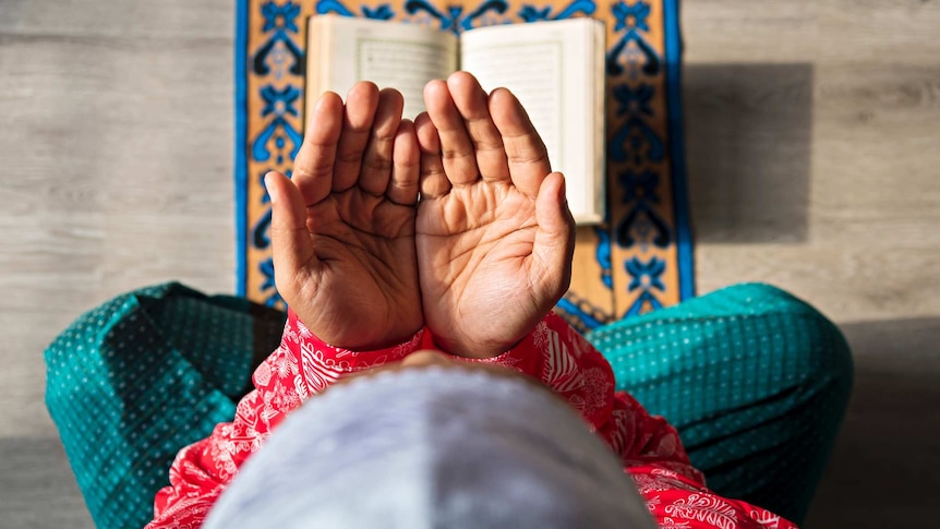 top view of man sitting crossed legged with hands stretched out above a sacred book placed on prayer mat