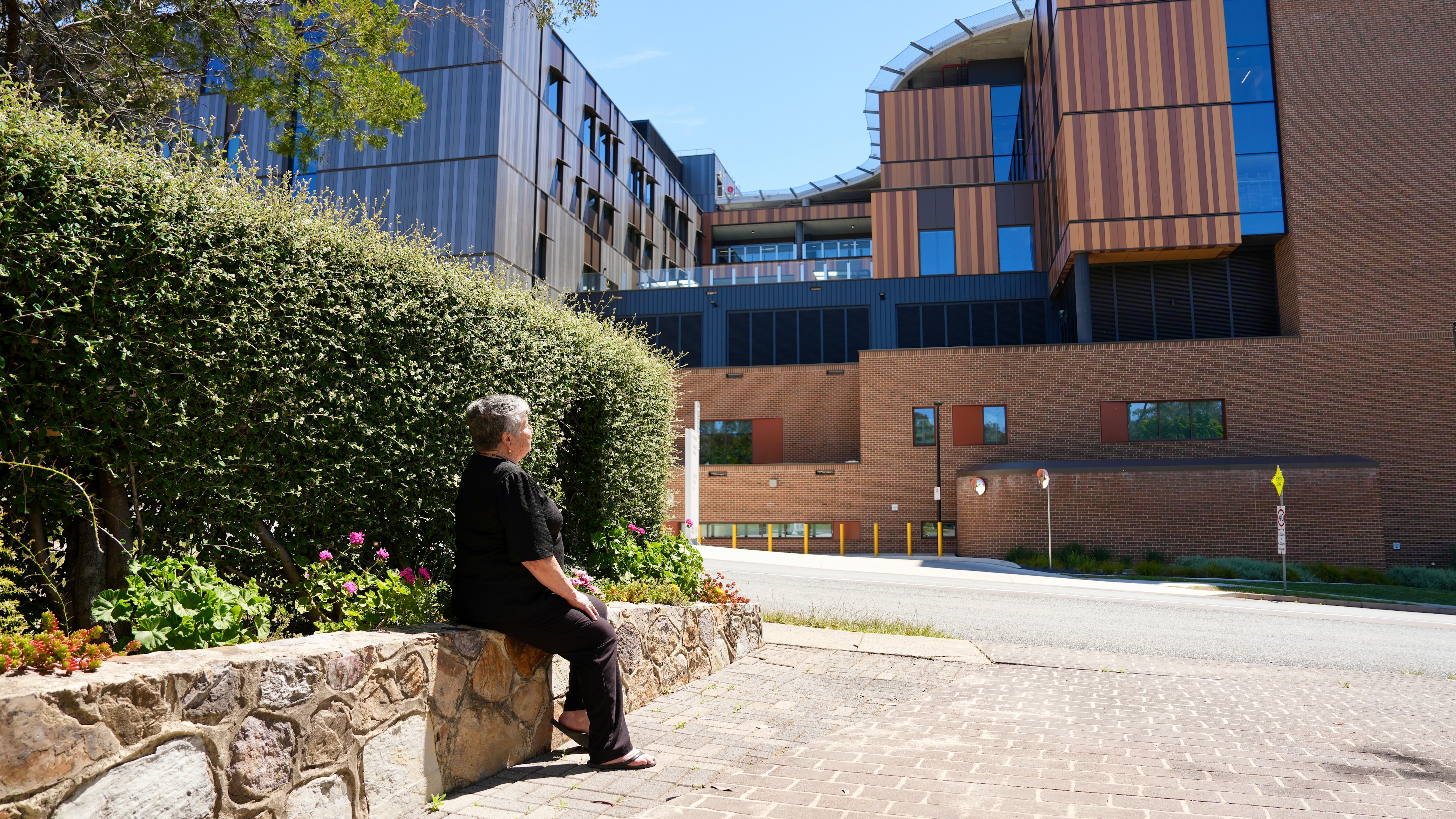 An 80-year-old woman with short grey hair, black blouse and necklace outside the hospital on a sunny day.