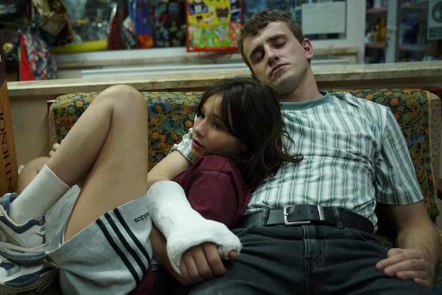 A 12 year old girl leaning on a man in his late 20s with an arm in a cast, sitting on a couch