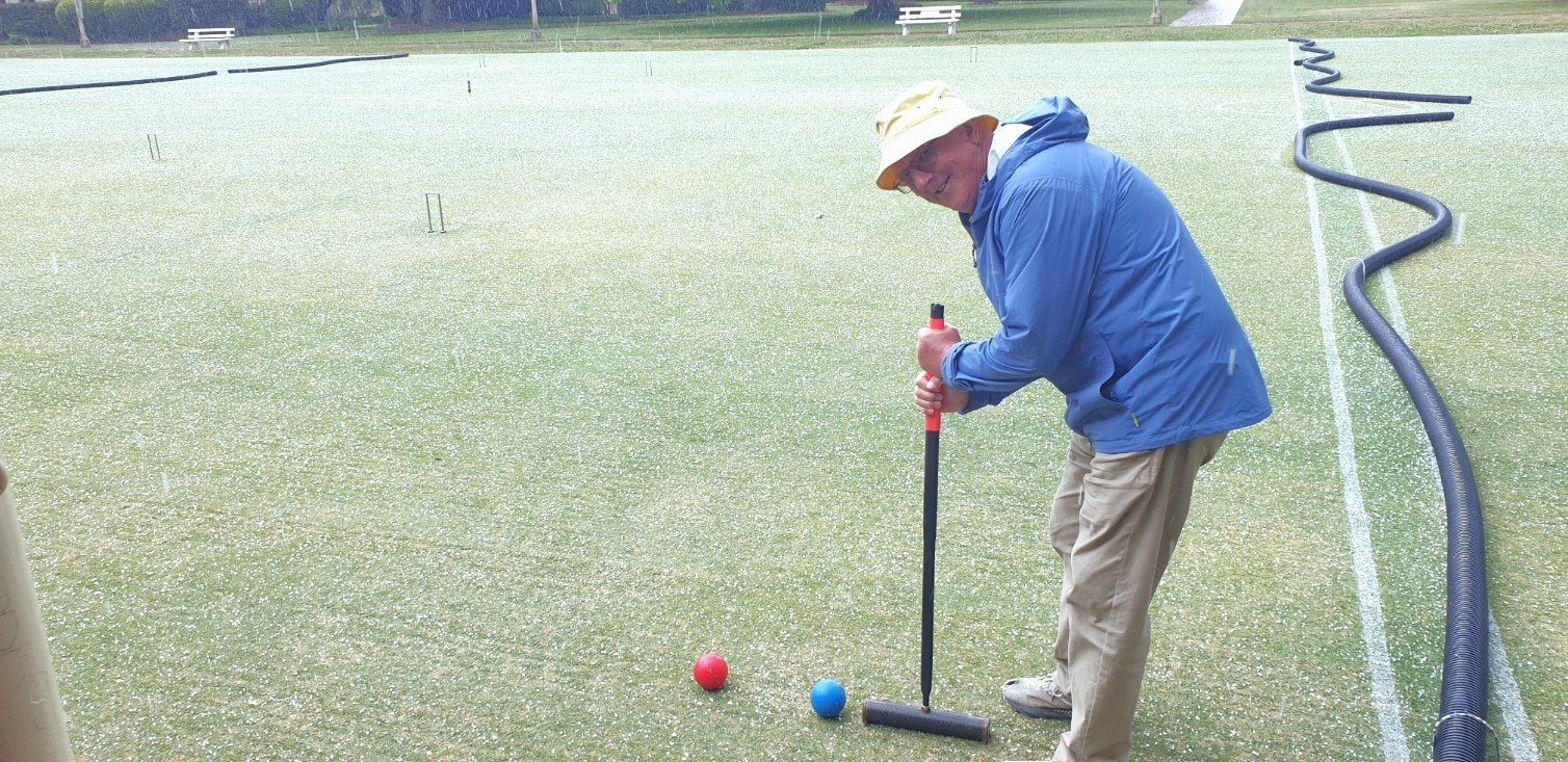 Homem prestes a acertar uma bola azul em uma bola vermelha usando um martelo de madeira de croquet, vestindo um casaco azul e um chapéu amarelo sorrindo para a câmera
