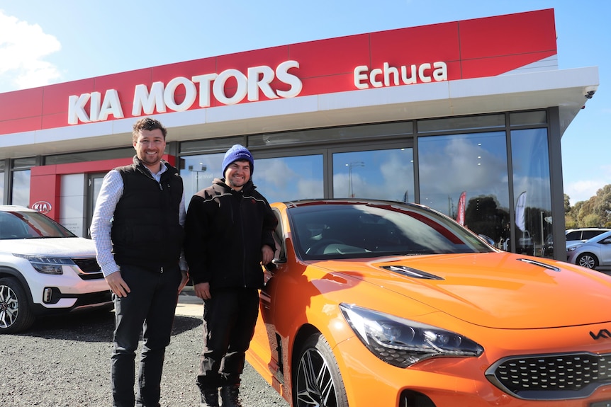 Two men smile in front of orange sports car parked outside building with red trim.