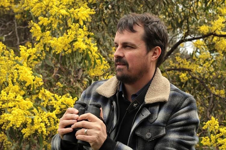 Sean Dooley with binoculars in hand stands in front of a yellow-flowering wattle tree