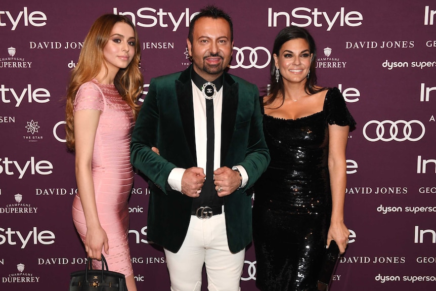 Napoleon Perdis, centre, wears an emerald blazer, black tie and white trousers as he is flanked by his wife and daughter.