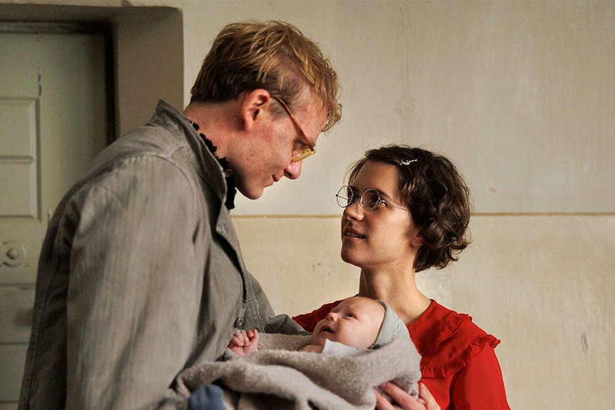 A film still of Johannes Hegemann and Liv Lisa Fries holding a baby between them. They're in 40s-style dress.