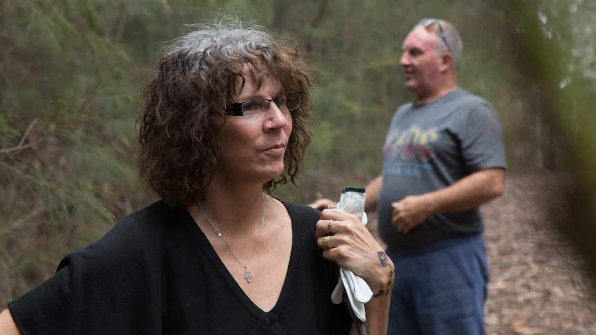 A woman holding gardening gloves and a stick pauses to think on a bush track