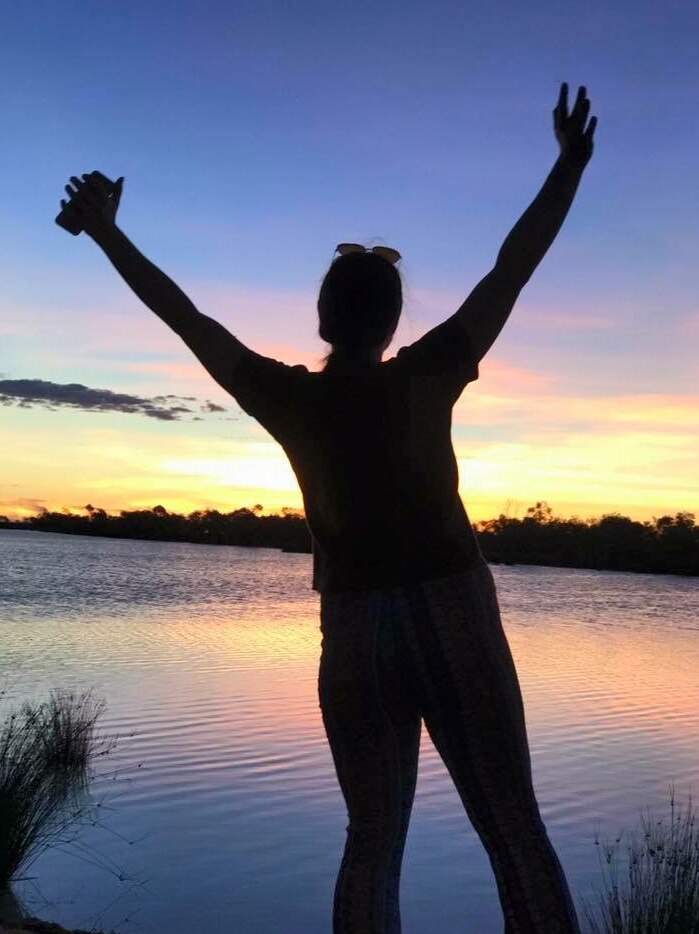 The dark silhouette of a woman with her hands in the air in front of the Pelican Point Billabong at sunset.