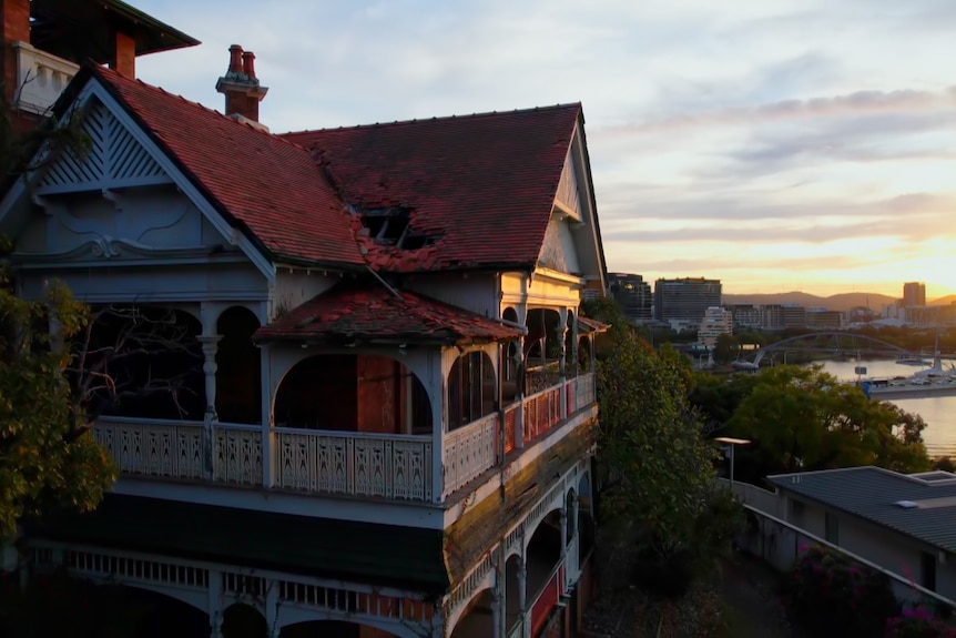 Una toma amplia de una antigua casa de la época de la federación a la que le faltan tejas.  El sol se pone detrás del río Brisbane a la derecha.