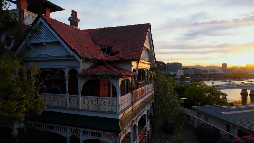 A wide shot of an old federation-era house with missing roof tiles. The sun is setting behind the Brisbane river to the right