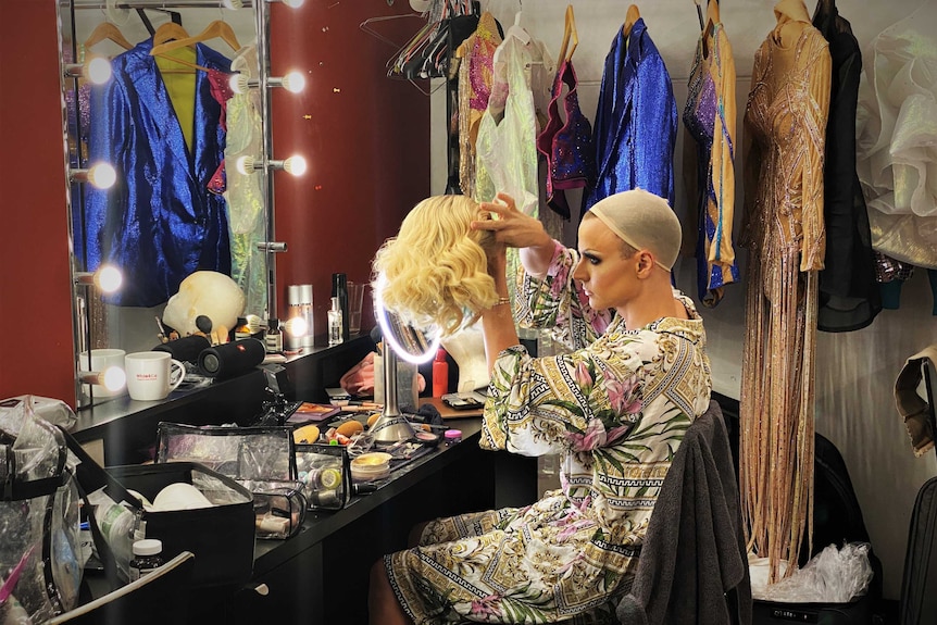 A man sitting on a chair wearing a floral robe, surrounded by costumes, holds a blonde wig