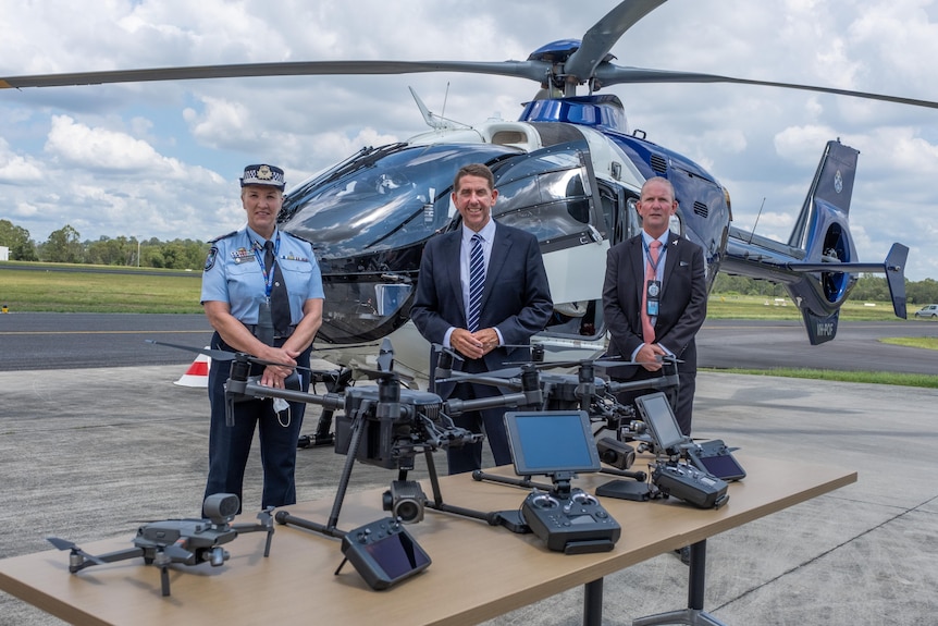 A policewoman and two men in suits stand near a police helicopter a table full of drones.