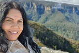 Kathleen has black hair and is smiling in a selfie. Behind her is a view of the luscious green valleys in the Blue mountains.