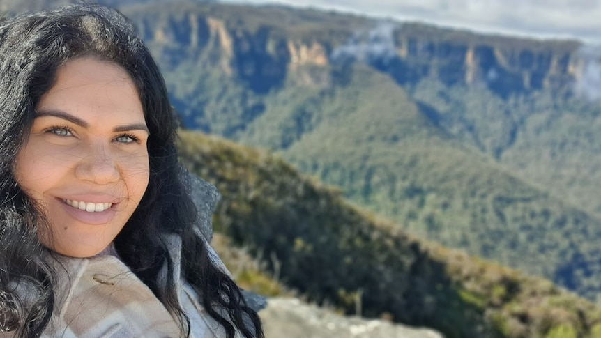 Kathleen has black hair and is smiling in a selfie. Behind her is a view of the luscious green valleys in the Blue mountains.