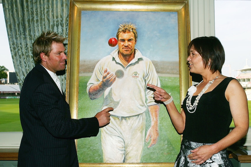 A woman points at a man, as they stand in front of a portrait of the man
