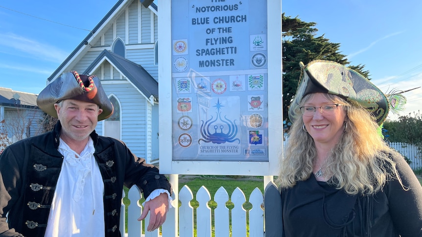 Two people wearing pirate hats stand near a sign for the "notorious" blue church of the flying spaghetti monster.