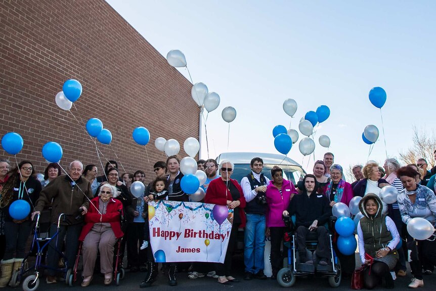 About 40 people holding blue and white balloons and a sign saying Happy Birthday James with a man in a wheelchair