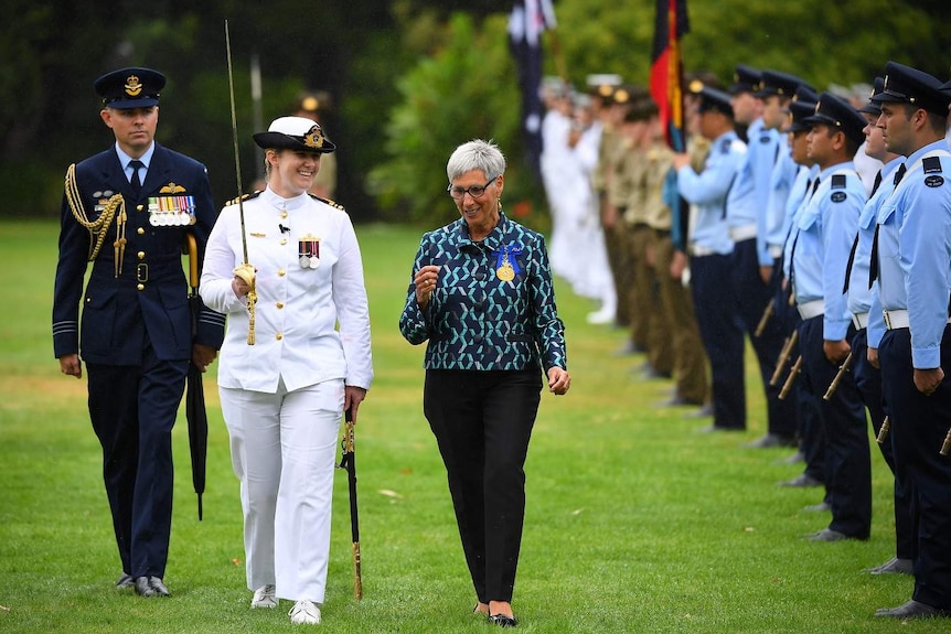 Governor Linda Dessau smiles as she walks past the military guard on a green lawn.