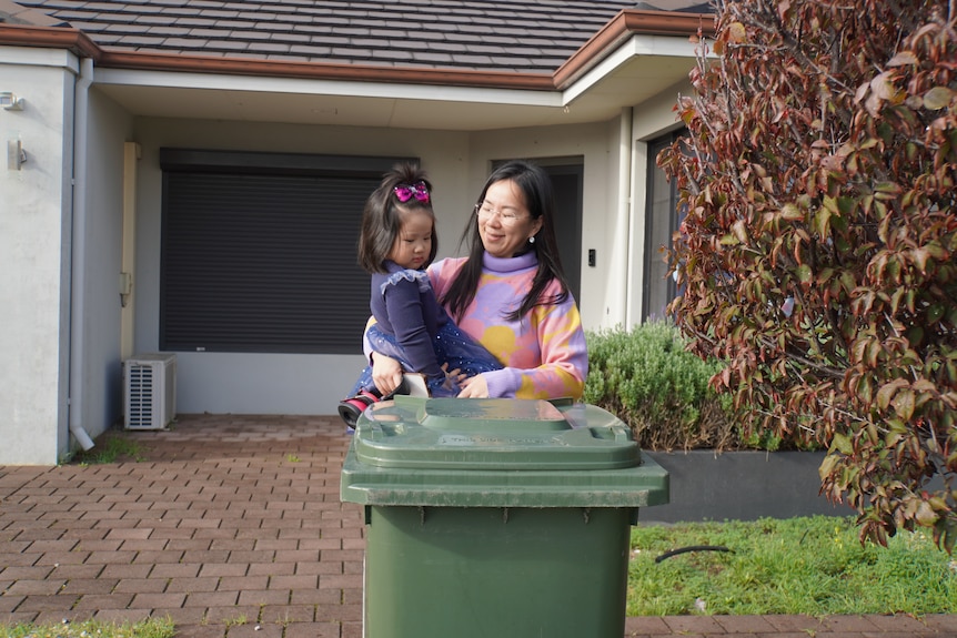 A woman stands outside a house carrying a young toddler in her arms behind a green bin.