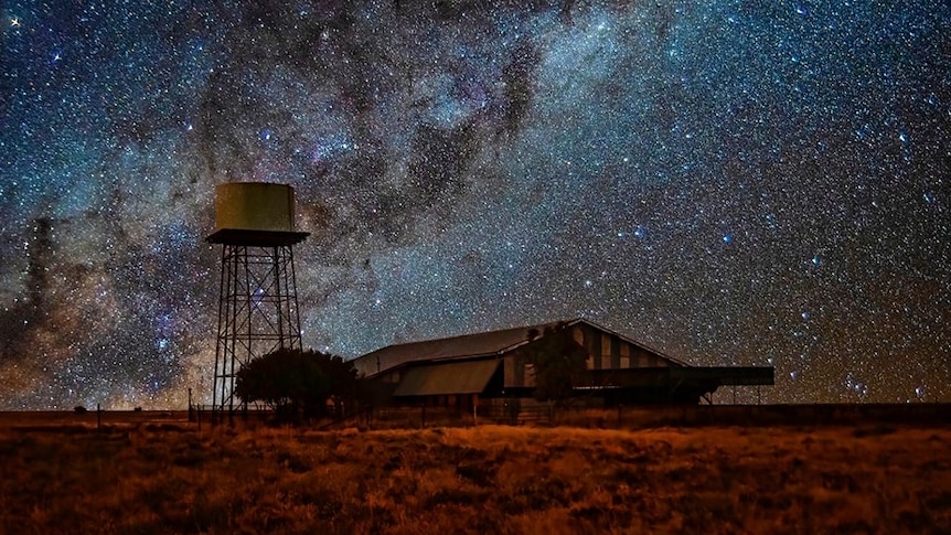 The Milky Way Galaxy of stars stretches across a dark night sky, with a large shed and water tank in the foreground.