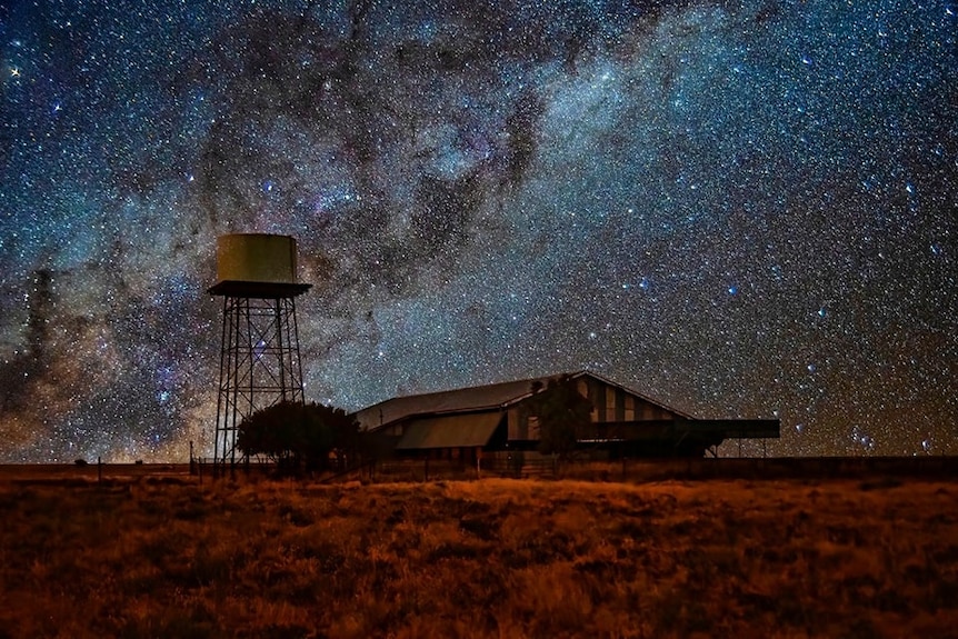 The Milky Way Galaxy of stars stretches across a dark night sky, with a large shed and water tank in the foreground.